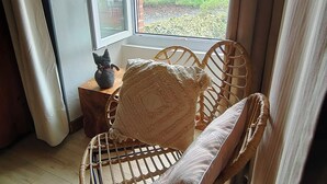 Uma cadeira de vime com almofada, uma mesinha com objeto decorativo, e vista para uma paisagem verde pela janela.
