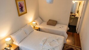 Habitación de hotel con dos camas, un ventilador de techo, un cuadro, una lámpara de noche, una mesita de noche y un baño visible a través de una puerta abierta.