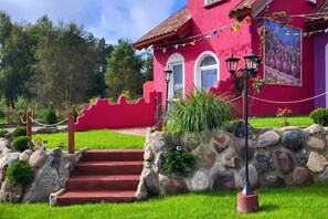 A pink building with a red roof, stone steps, and a garden with grass and plants.