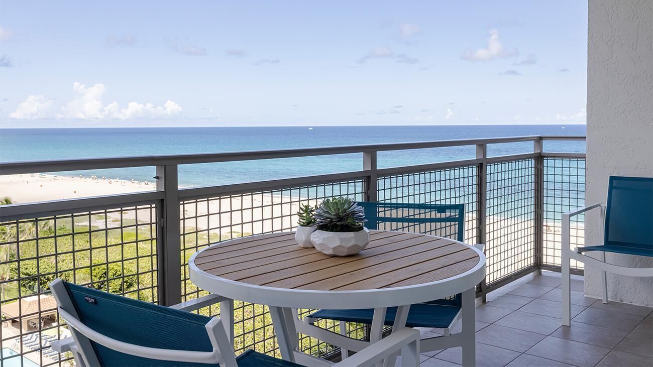 A balcony with a view of the beach, a round table with chairs, and a potted plant.