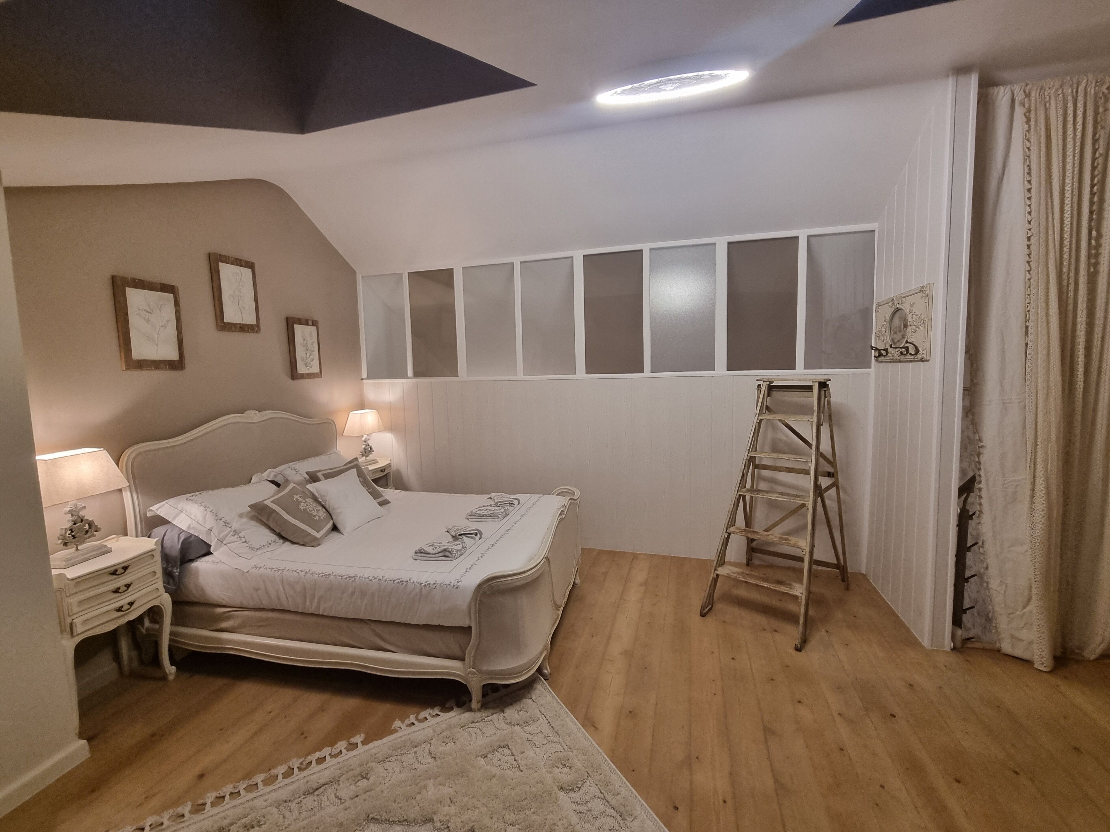 Un dormitorio con cama, mesitas de noche, una escalera, una ventana con persianas y una lámpara en el techo.