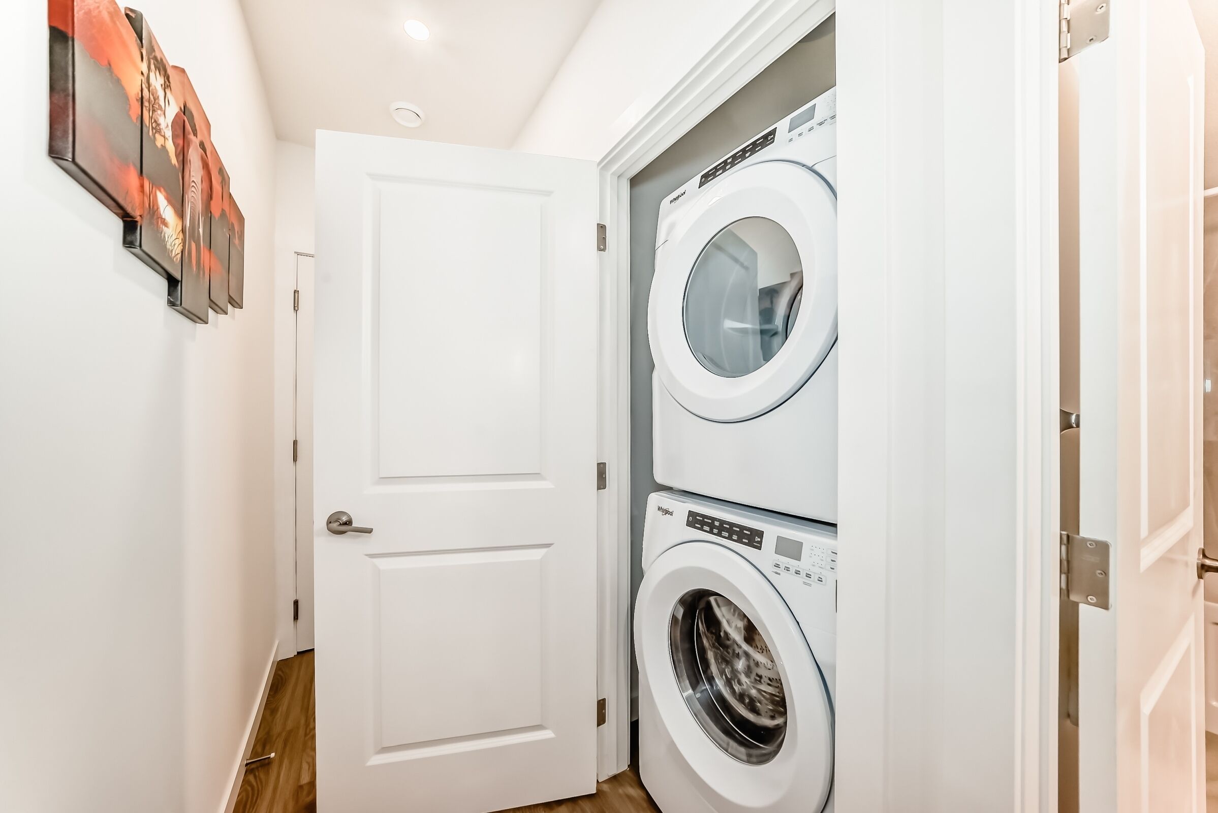 A laundry room with two stacked washing machines, a white door, and a hallway.