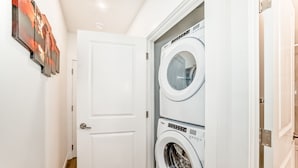 Uma lavanderia com duas máquinas de lavar empilhadas, uma porta branca e um corredor.