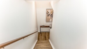 Uma escadaria estreita com degraus de madeira e corrimão, que leva a um patamar com uma pequena mesa e uma obra de arte emoldurada.