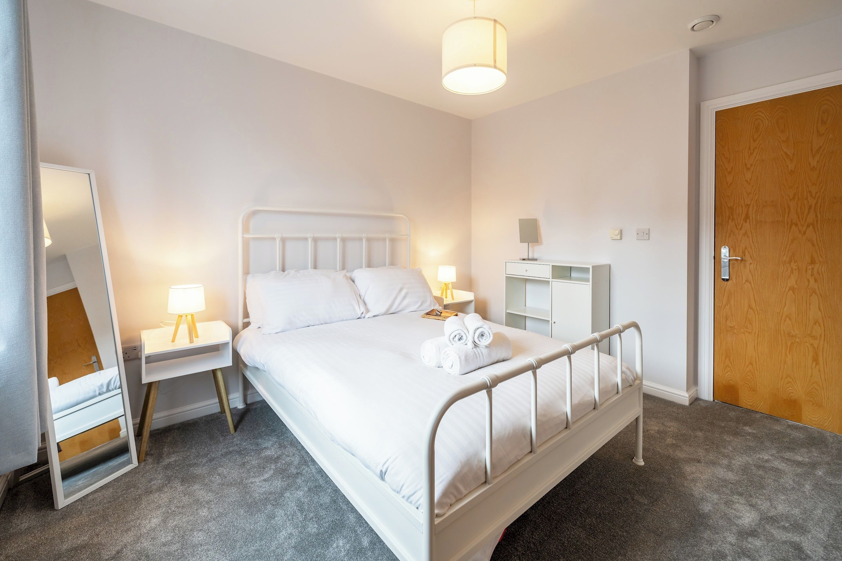 A neatly arranged bedroom with a bed, bedside tables, a mirror, and a wooden door.