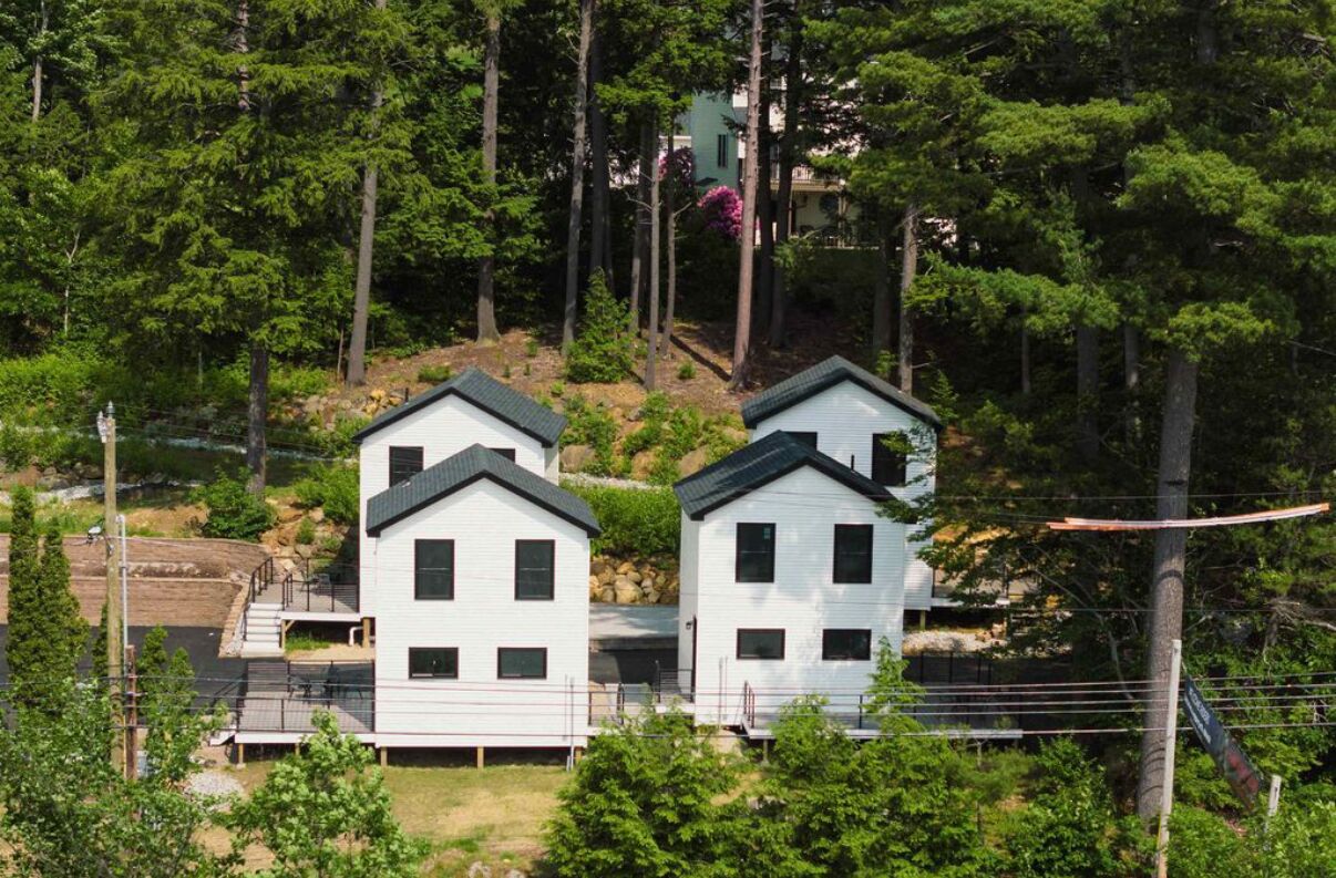Two white houses with dark roofs, surrounded by dense green trees and a rocky hillside.