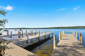 Un muelle de madera se extiende hacia un lago tranquilo con aguas cristalinas, rodeado de vegetación y una orilla distante.