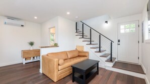 A modern living room with a brown leather sofa, a black coffee table, and a wooden console table. A staircase with a black railing leads to a white door. - Lakeside Cottage Retreat – 4 Loft Style Cottages with Dock Slip - Weirs Beach (Laconia)