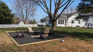 Un patio trasero con un pozo de fuego, sillas Adirondack y una casa blanca al fondo.
