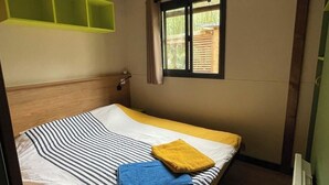 Ein Einzelbett mit gestreifter Bettdecke und einem gelben Kopfkissen, ein Fenster mit Blick ins Grüne und eine grüne Regalwand.