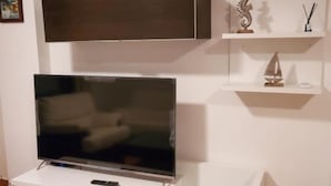 A living room with a flat-screen TV on a white stand, a glass-top coffee table, and a white sofa. - Charming Apartment With Garden in Almeria (Vera)