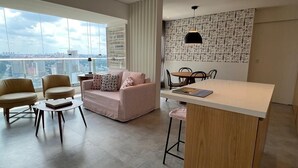 A modern living room with a sofa, chairs, a round table, a kitchen area with a bar, and a city view through large windows. - Best View in SP - 62 m2 Apartment - 10 min from Butantã Metro Station (Yellow Line) (São Paulo)