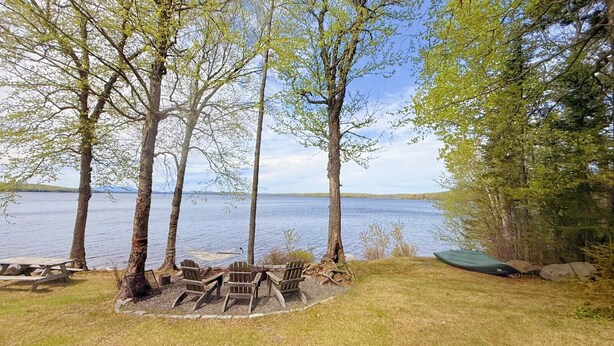 A lakeside camping area with a picnic table, four Adirondack chairs, and a canoe.