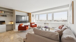 A modern living room with a large TV, red and beige sofas, a coffee table, and a city view through large windows. - 2-Bed Apartment with Views of Plaza de Colón (Madrid)