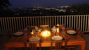 Mesa de jantar posta para seis pessoas, com copos de vinho, pratos e uma vela, com vista para um horizonte urbano à noite.