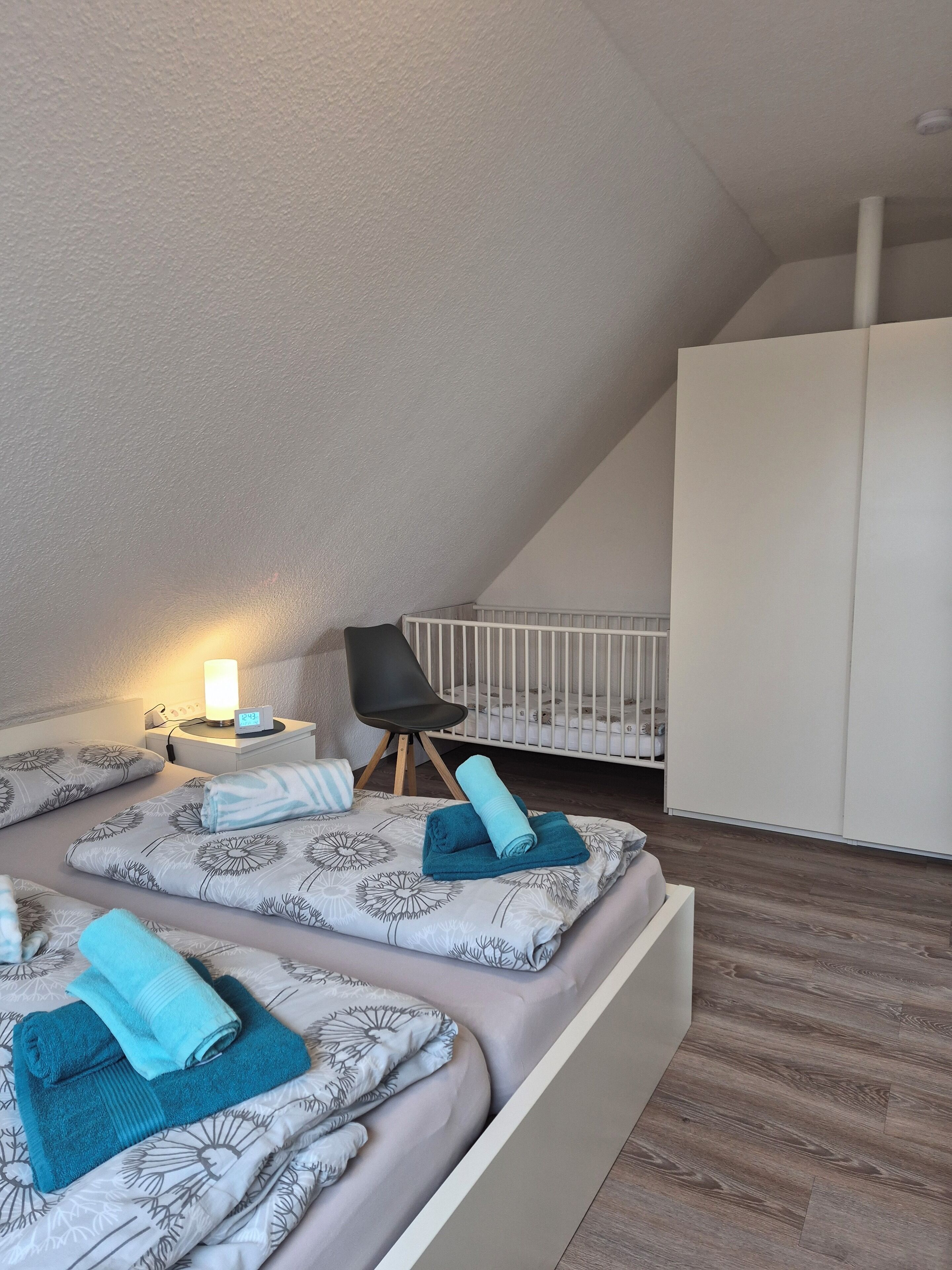 Un dormitorio con techo inclinado, una cuna, una mesita de noche con lámpara y una silla.