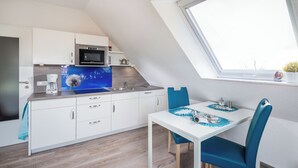 A compact kitchen with white cabinets, a microwave, and a coffee maker. There's a dining table with blue chairs and a view of the sky through a skylight. - Vacation apartment "Tillmanns Pusteblume" in Norddeich - quiet & close to the beach (Norden)