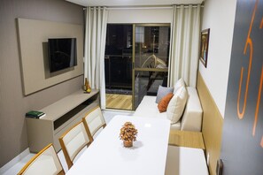 Una camera d'albergo moderna con TV a schermo piatto, un tavolo da pranzo con sedie, un divano con cuscini e un'ampia finestra con tende.