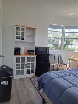 A compact room with a bed, a small kitchenette, a window with a view of palm trees, and a trash bin. - Phenomenal Studio - Steps to Beach - Great Views - Historic - terrific stay (Surfside)