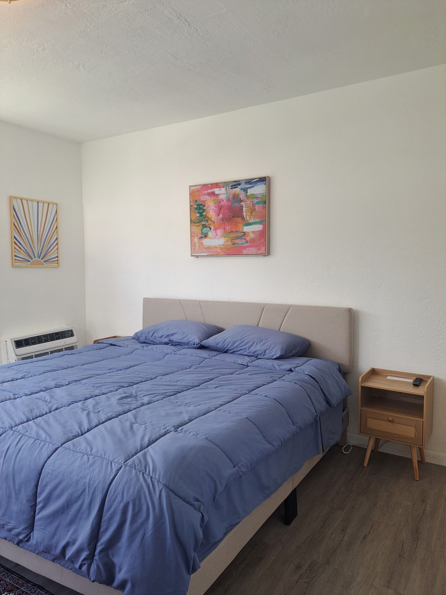 Ein Schlafzimmer mit einem Bett, einem Nachttisch, einer Wand-Klimaanlage und einem farbenfrohen abstrakten Gemälde.