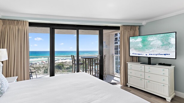 Ein Hotelzimmer mit einem großen Bett, einem Fernseher, einem Balkon mit Blick auf den Strand und einer weißen Kommode.