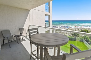 Un balcon avec une table et des chaises, donnant sur une plage oĂč lâon peut voir des gens et des parasols.