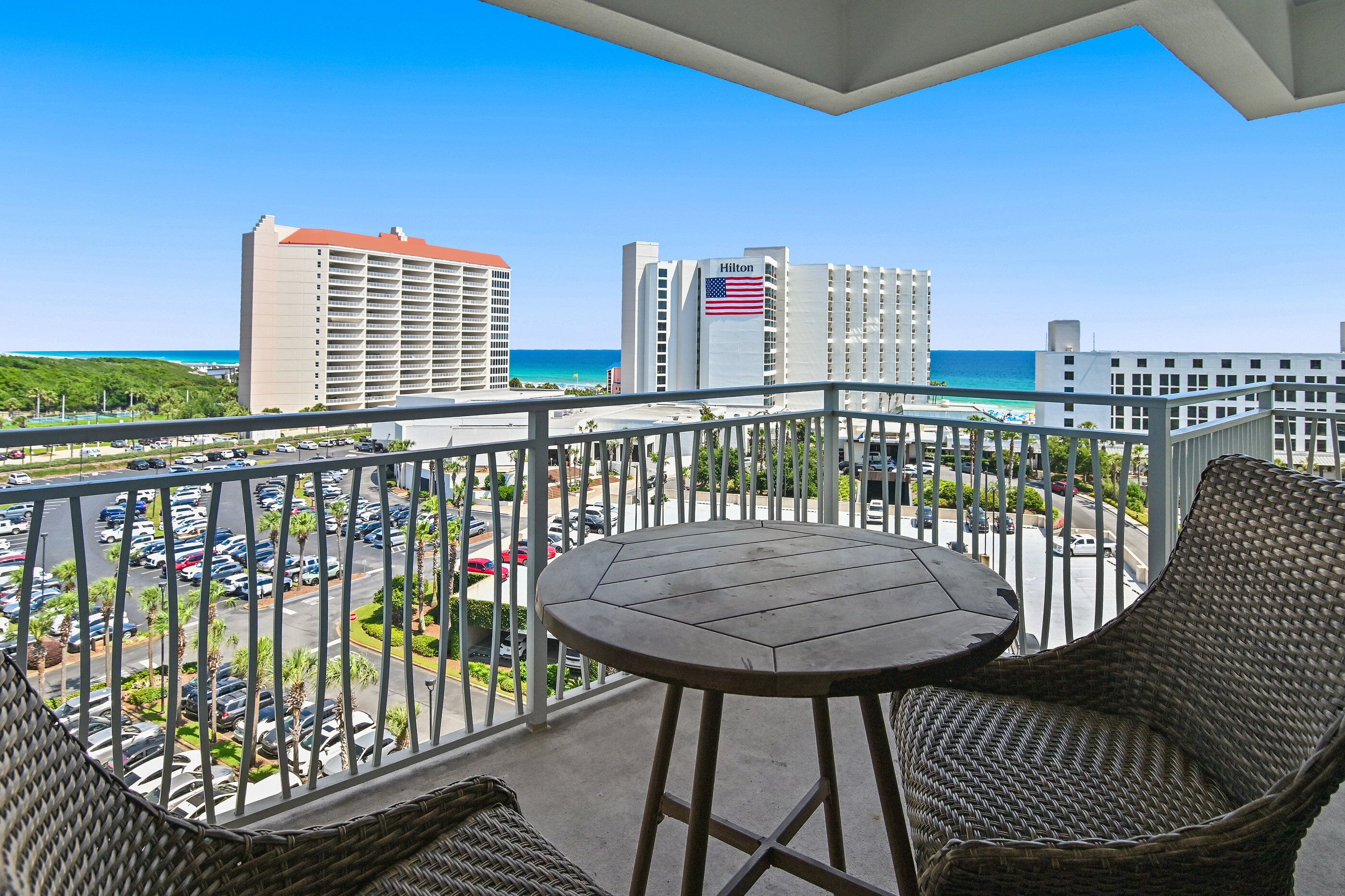 A balcony with wicker chairs and a table, overlooking a parking lot and buildings with an American flag.