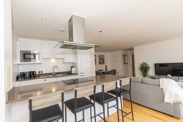 A modern kitchen with a central island, bar stools, a grey sofa, and a flat-screen TV in a living area. - Greenwich Apartment Near Cutty Sark and Paid Parking (London)