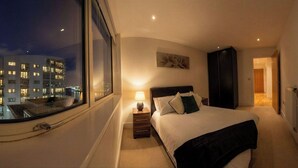 A hotel room with a large window, a bed, a bedside table with a lamp, a dark headboard, and a door leading to another room. - Greenwich Apartment Near Cutty Sark and Paid Parking (London)