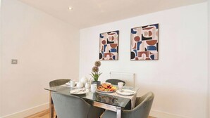 A dining area with a glass table, grey chairs, a white radiator, and abstract wall art. - Greenwich Apartment Near Cutty Sark and Paid Parking (London)