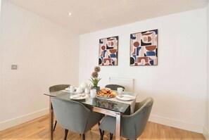 A dining area with a glass table, grey chairs, a white radiator, and abstract wall art. - Greenwich Apartment Near Cutty Sark and Paid Parking (London)