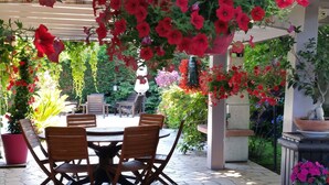 Un espace patio avec une table ronde et des chaises, entouré de plantes en pot et de fleurs suspendues.