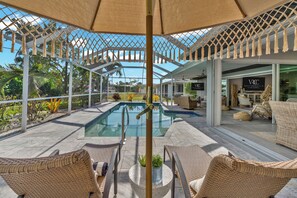A poolside area with wicker chairs, a glass pool, and a covered seating area with a sign for VRCC Vacation Rentals. - VRCC Villa Cozy Cape (Cape Coral)