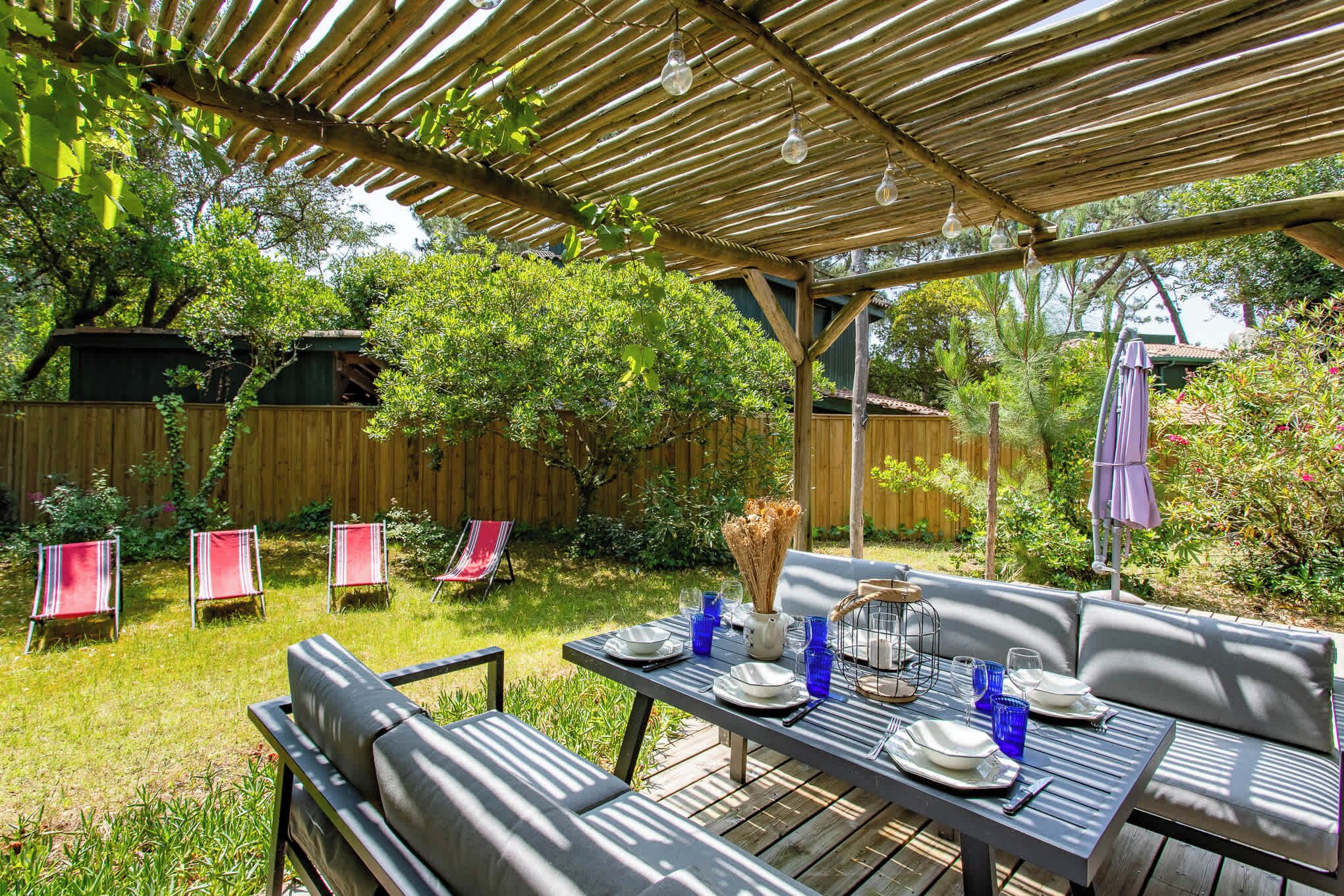A covered patio with a table set for dining, surrounded by greenery and a wooden fence.