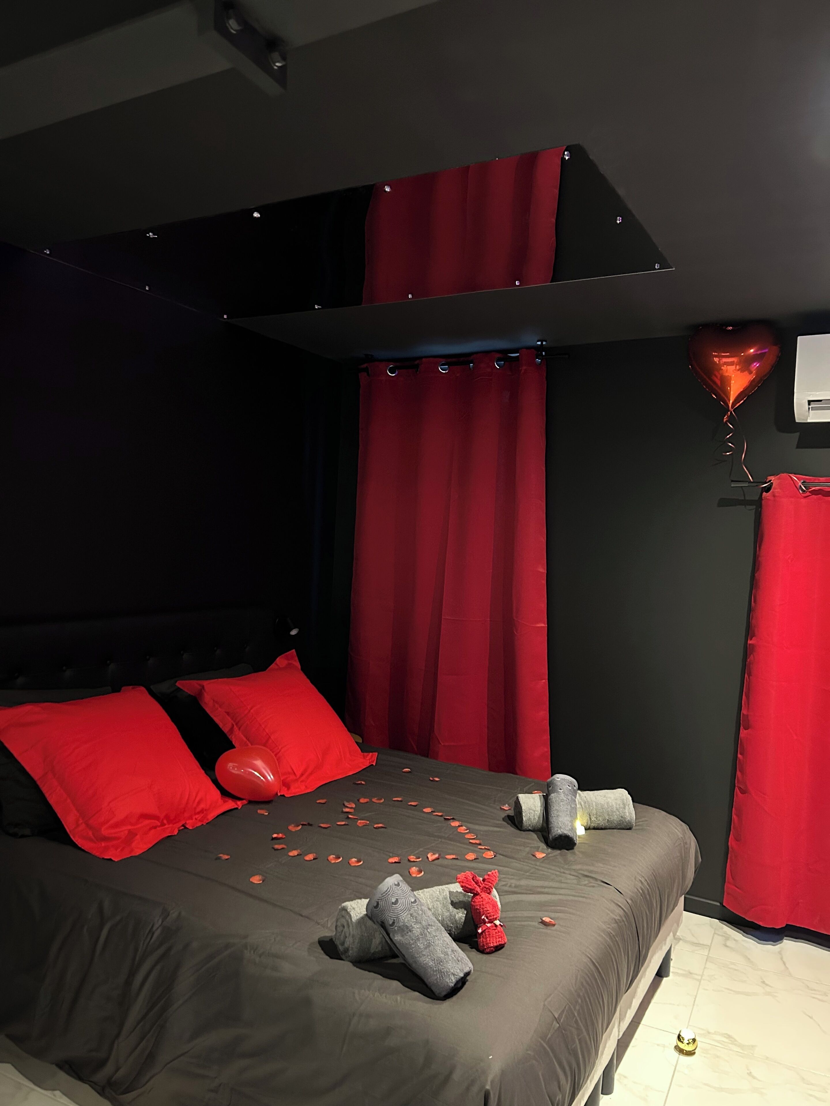 A bed with red pillows, a heart-shaped balloon, and rose petals on the floor.