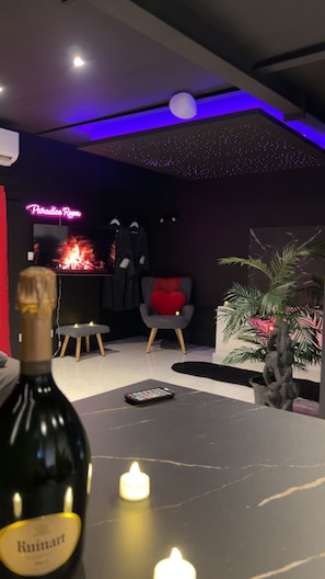 Ein Zimmer mit einer Neonreklame, einem Kamin, einem Sessel mit rotem Kissen, einer Weinflasche und Kerzen.