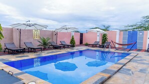 Une piscine entourée de chaises longues et de parasols, bordée d’un mur rose et dotée d’un sol en pierre.