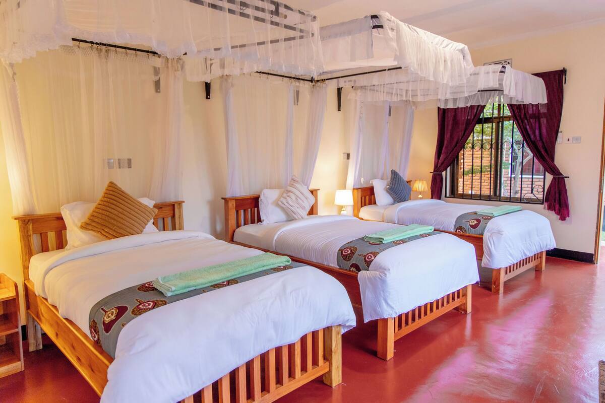 Une chambre avec trois lits en bois, du linge de lit blanc, une moustiquaire et une fenêtre avec des rideaux.