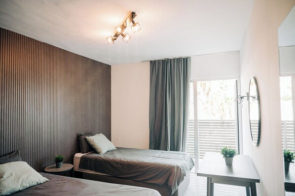 A modern bedroom with two beds, a small table, a potted plant, and a large window with blinds. - Napa Rocks Superiors  (Ayia Napa)