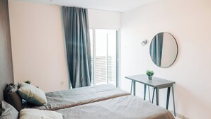Quarto com duas camas, uma mesa de cabeceira com uma planta, um espelho redondo na parede e uma janela com cortinas.