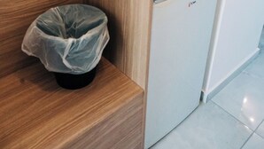 Canto de um quarto com um banco de madeira, uma pequena lixeira e uma geladeira compacta.