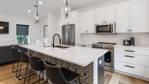 Una cucina moderna con mobili bianchi, elettrodomestici in acciaio inossidabile, una grande isola con sgabelli da bar e luci a sospensione.