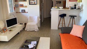 En kompakt opholdsstue med et fjernsyn, en stol med ryglæn, et spiseområde med barstole og et køkken i baggrunden.