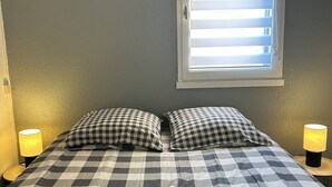 Ein Schlafzimmer mit kariertem Bettbezug, zwei Kissen, einem Fenster mit Jalousien, einer Lampe auf jeder Seite und einem zusammengerollten Handtuch auf dem Bett.