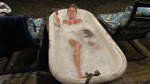 Een bad vol water, iemand zit erin, en een stoel met een decoratieve bekleding.