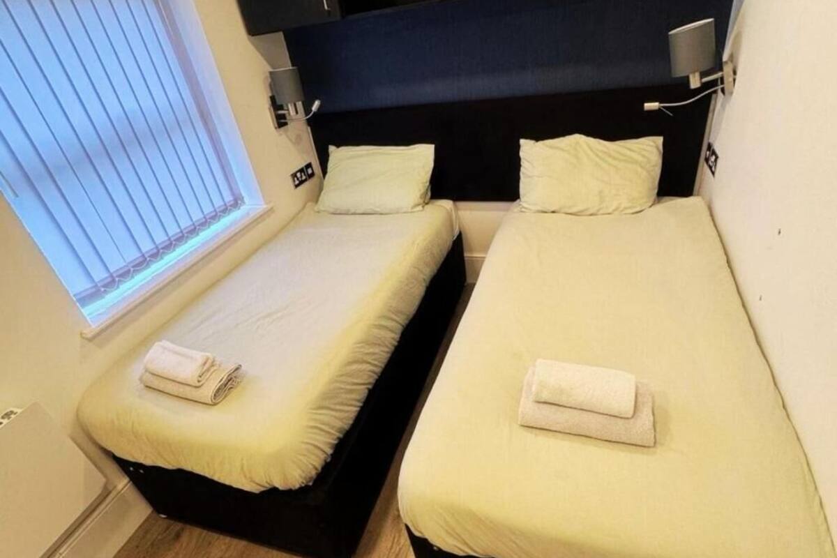 Deux lits simples avec du linge de lit blanc, une fenêtre avec des stores et une lampe fixée au mur.