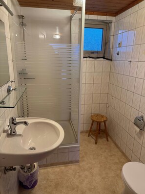 Ein Badezimmer mit Duschkabine, Waschbecken mit Chromarmatur, Toilette und einem kleinen Hocker.