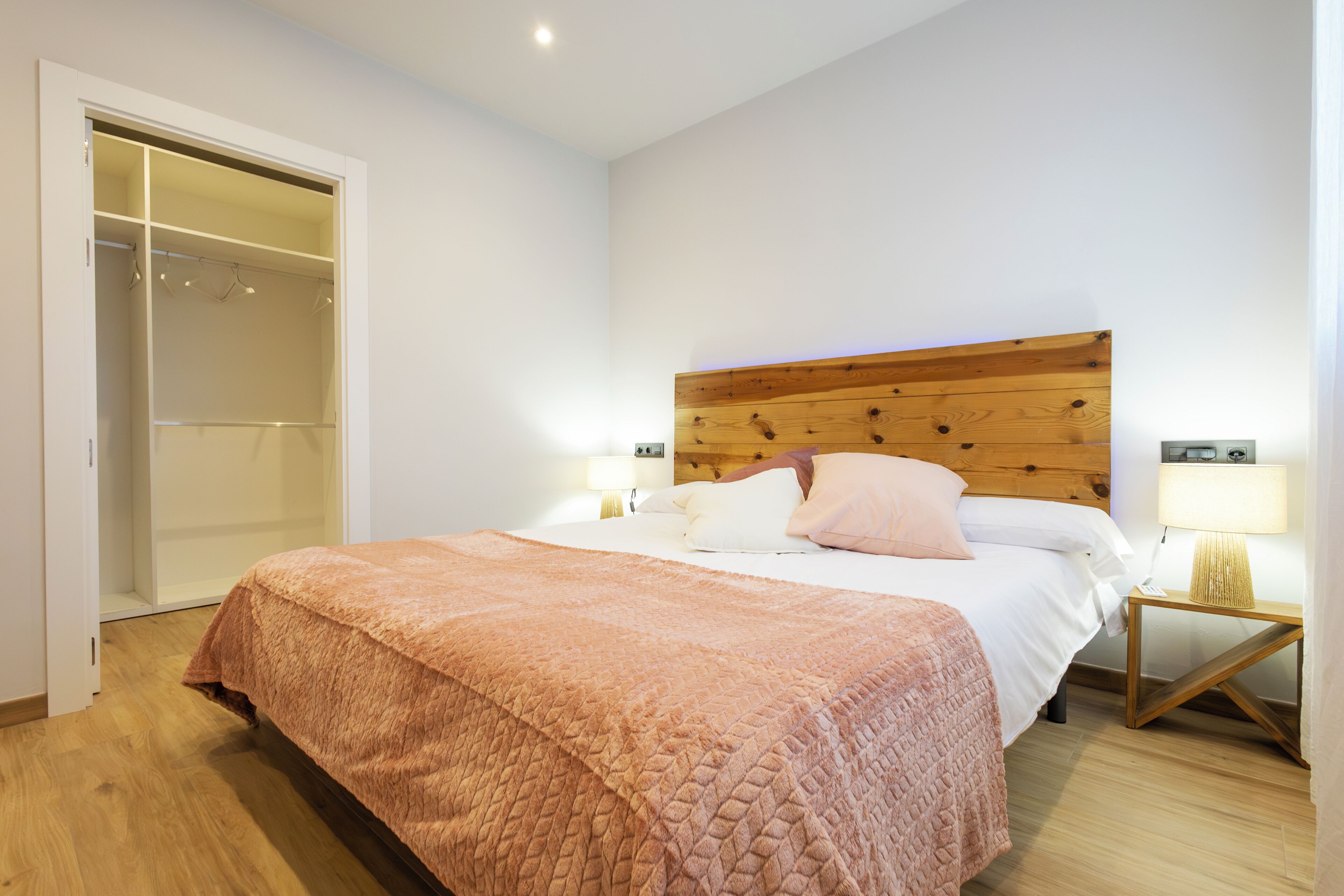 A bedroom with a wooden headboard, a bed with white linens, a pink blanket, a wooden nightstand with a lamp, and a walk-in closet.