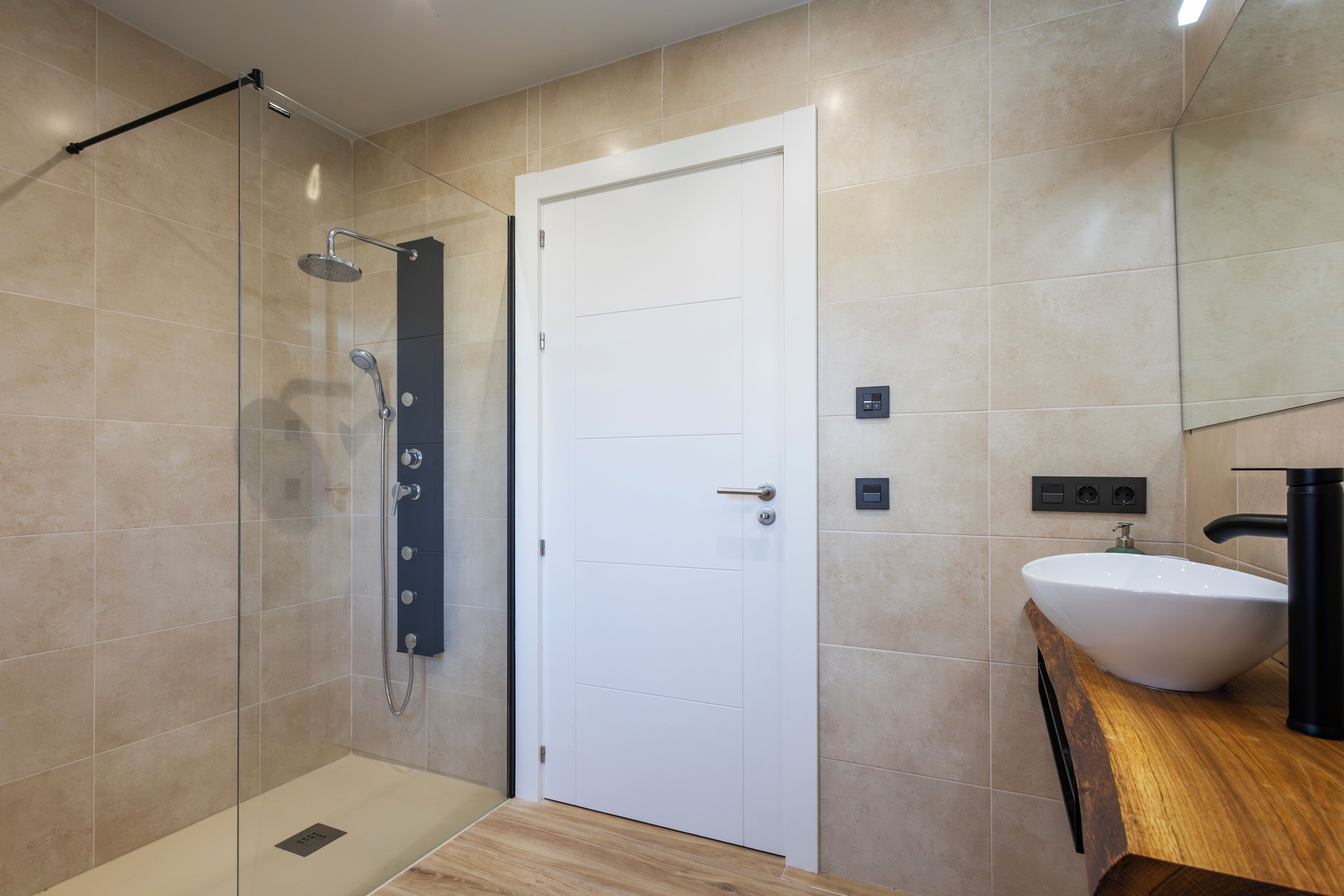 Un baño moderno con cabina de ducha de vidrio, una puerta blanca y un tocador de madera con lavamanos.
