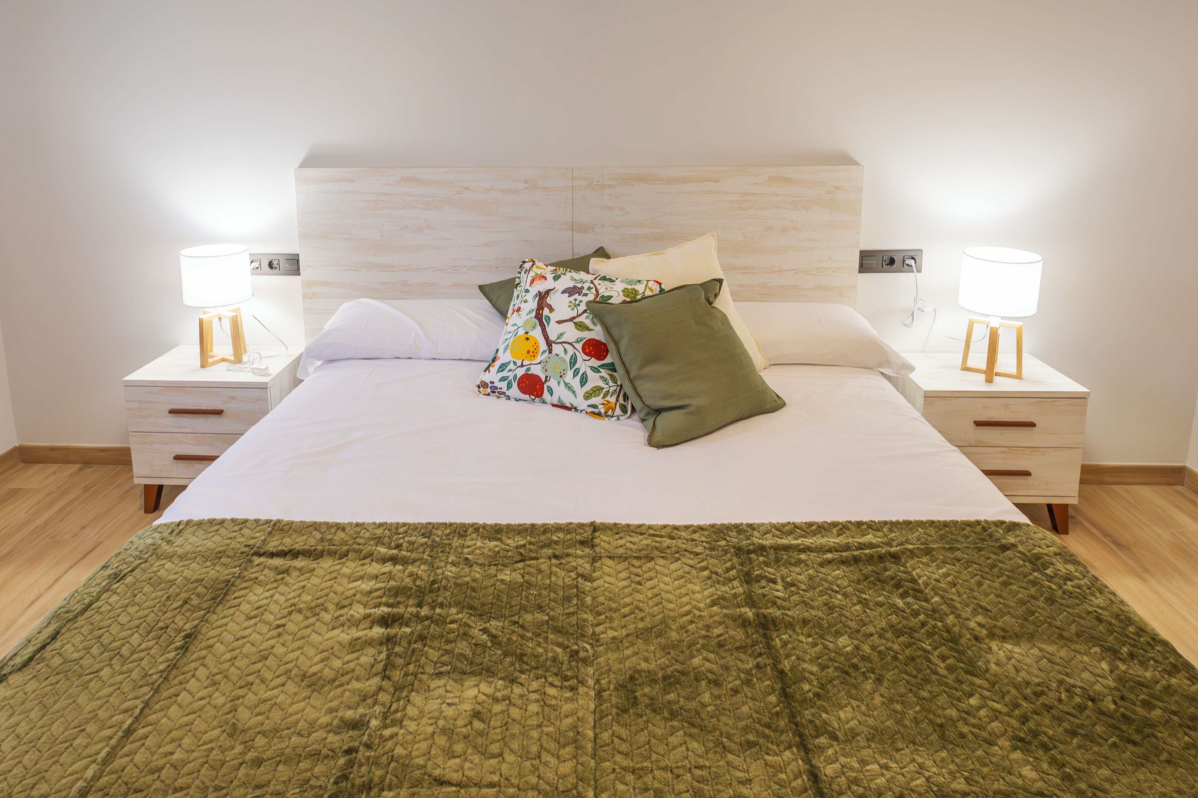 A neatly made bed with a green blanket, two bedside tables with lamps, and a wooden headboard.